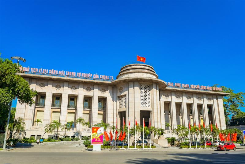 A grand government building with a curved central tower displays a Vietnamese flag, surrounded by flags and palm trees, under a clear blue sky.